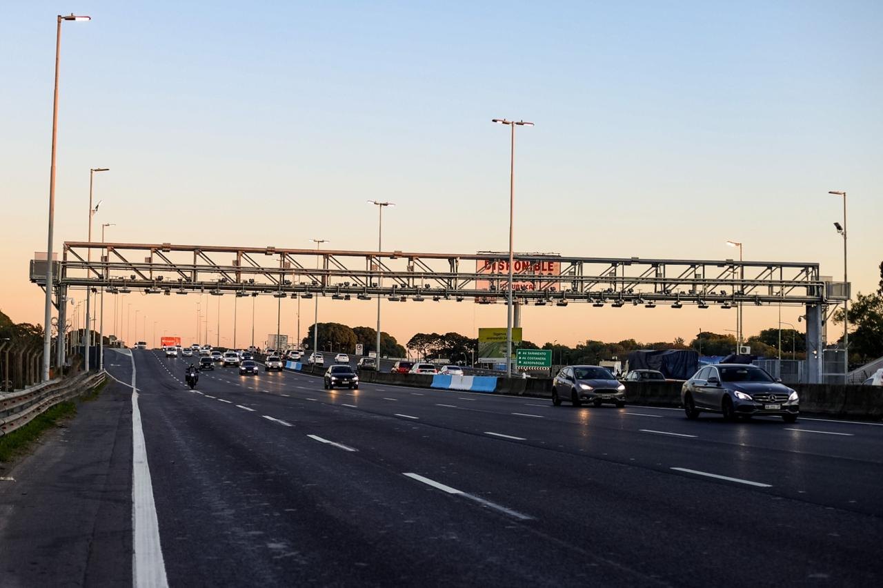 Autos circulando por la Autopista Perito Moreno (AU Illia) en CABA al atardecer, bajo un pórtico de peaje, con una moto al fondo.